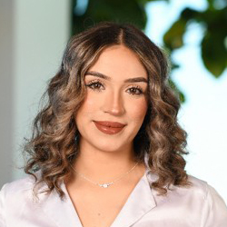 A young woman with shoulder-length, wavy brown hair wearing a white collared shirt and a simple silver necklace.